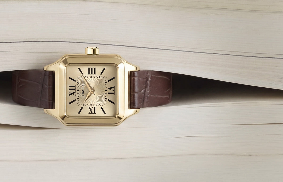 Timex Addison watch with a gold-tone rectangular case, champagne dial, black Roman numerals, and brown leather strap displayed on an open book.