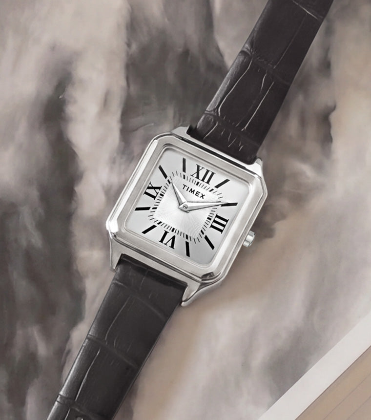 Timex Addison watch with a silver-tone rectangular case, white dial, Roman numerals, and black leather strap displayed on an open book.