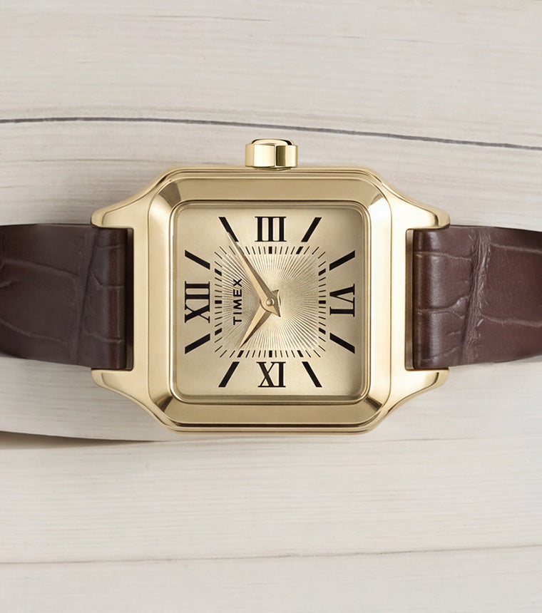 Timex Addison watch with a gold-tone rectangular case, champagne dial, black Roman numerals, and brown leather strap displayed on an open book.