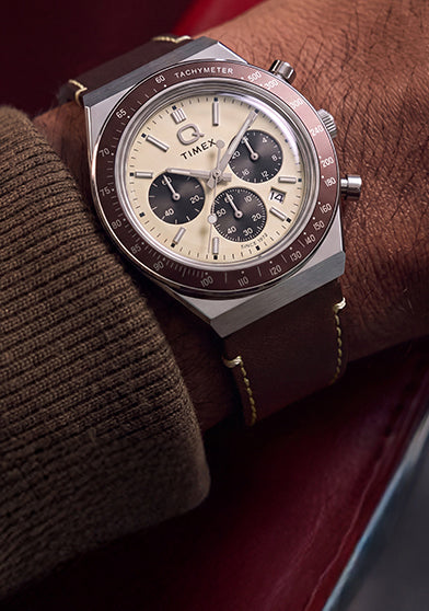 Q Timex Chronograph with a cream dial and brown leather strap styled on the wrist inside a red car interior.