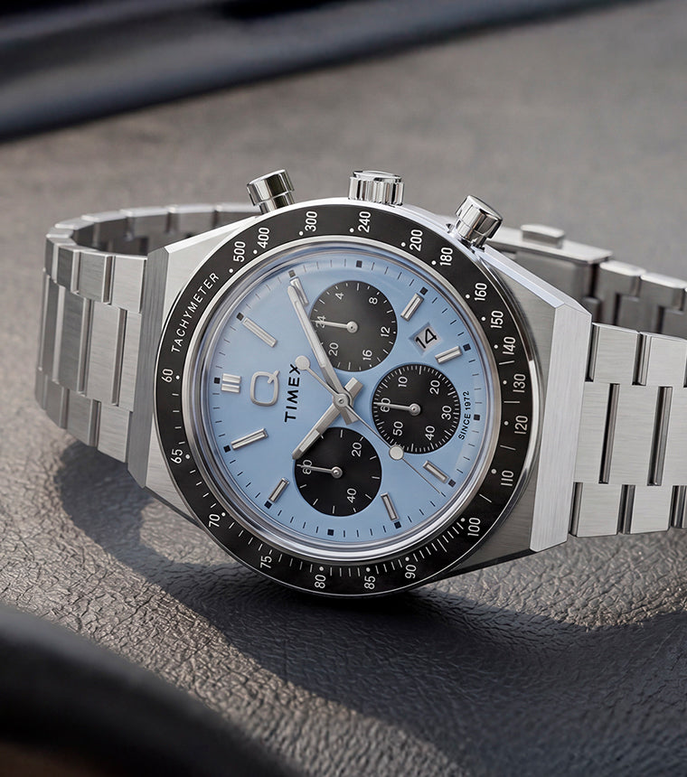 Q Timex Chronograph with an ice-blue dial and stainless-steel bracelet displayed on a black leather surface.