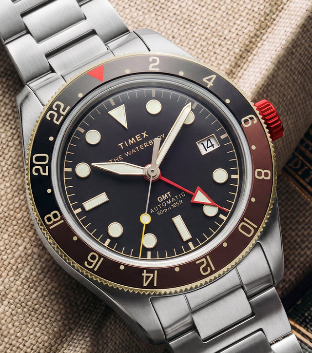 Waterbury Heritage Automatic GMT watch with a black dial, brown 24-hour bezel, red crown, and stainless-steel bracelet displayed beside vintage books.