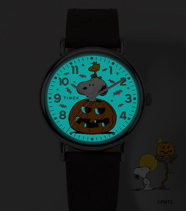 Peanuts® x Timex Weekender® Pumpkin watch shown in the dark with a glowing blue INDIGLO® dial revealing a bat pattern around Snoopy and Woodstock.