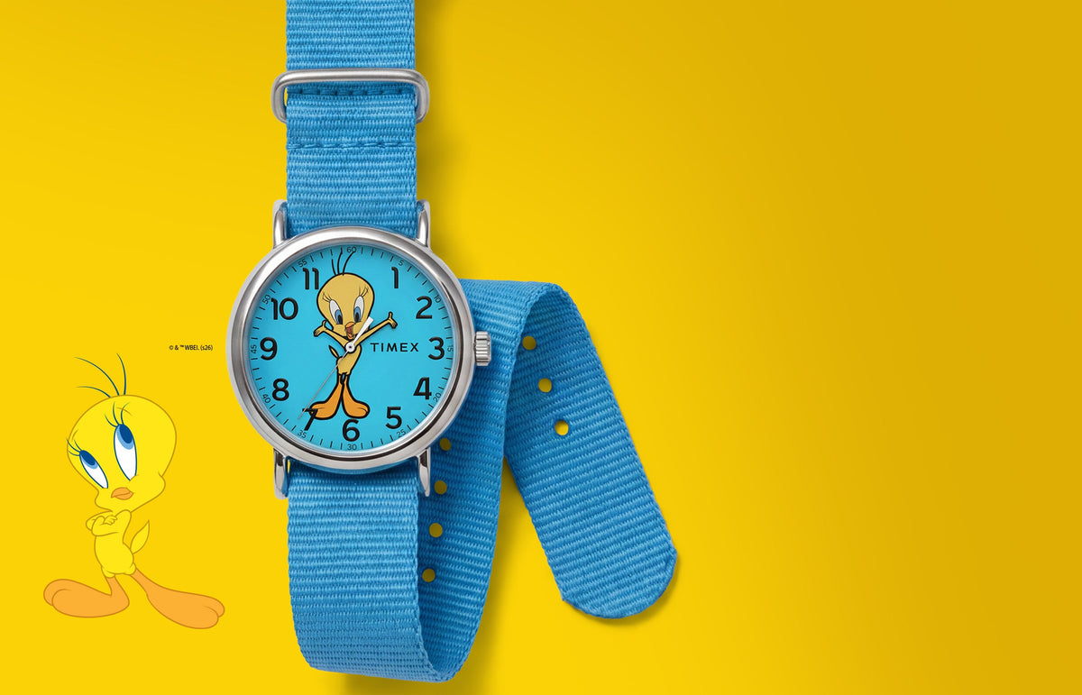 TW2Y99500 Looney Tunes Tweety Bird x Timex Weekender watch with a blue character dial and blue fabric strap beside Tweety artwork on yellow.