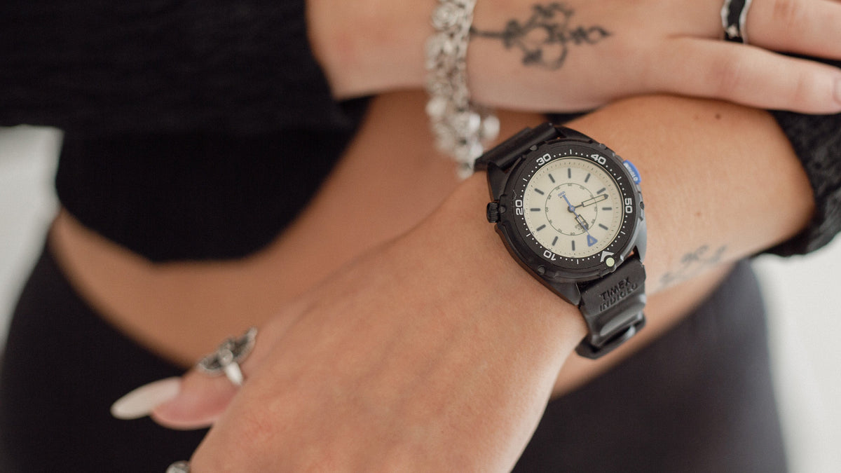 TW6A19800 Timex Intrepid x Dimepiece ‘Blackout’ Edition with a cream dial and black strap, worn on a wrist beneath a hand with silver jewelry.