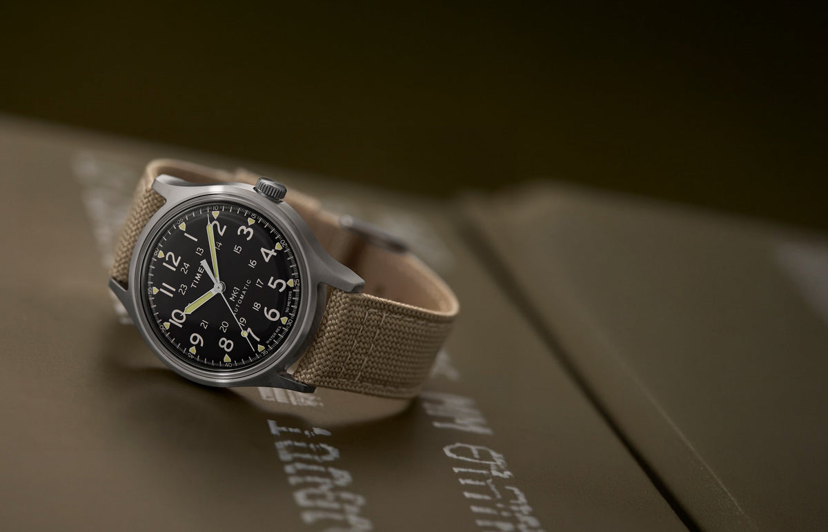 MK1® Automatic watch with a black dial, stainless-steel case, and tan fabric strap resting on military-style cases.