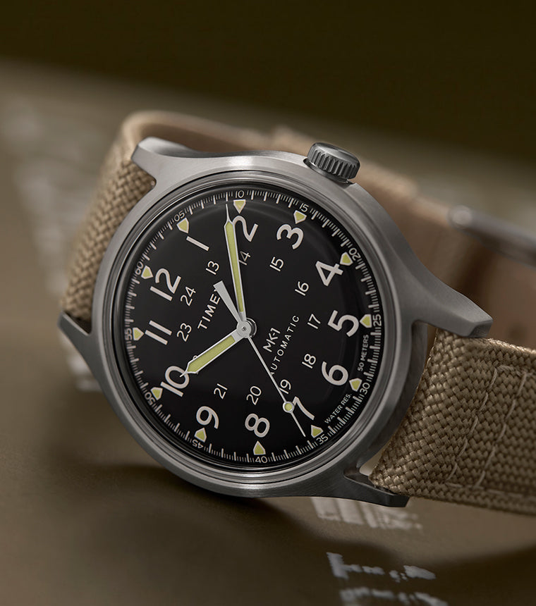 MK1® Automatic watch with a black dial, stainless-steel case, and tan fabric strap resting on military-style cases.
