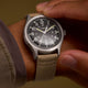 Close-up of the MK1® Automatic watch worn on the wrist, featuring a black dial and tan fabric strap.