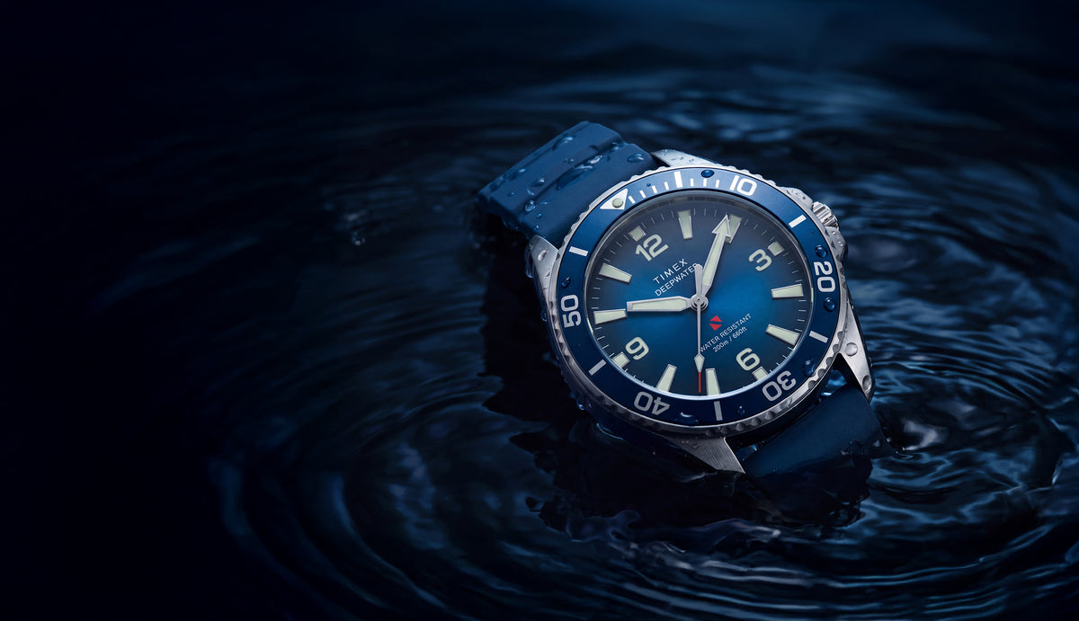 TW3A01900 Deepwater Arctic 200 watch with a blue dial, rotating bezel, and blue rubber strap resting in dark water.