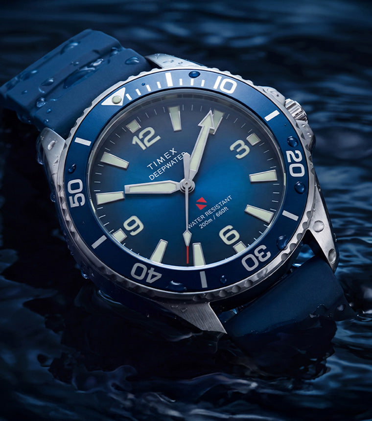 TW3A01900 Deepwater Arctic 200 watch with a blue dial, rotating bezel, and blue rubber strap resting in dark water.