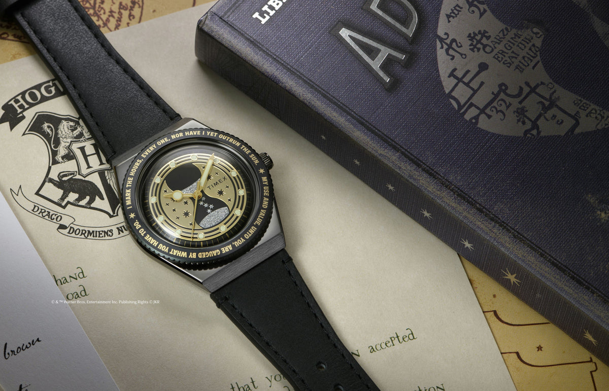 TW2Y76000 Black watch with a gold hourglass and stars on the dial, laid across Hogwarts-themed papers beside a book.