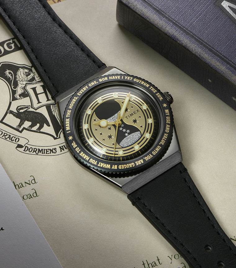 TW2Y76000 Black watch with a gold hourglass and stars on the dial, laid across Hogwarts-themed papers beside a book.