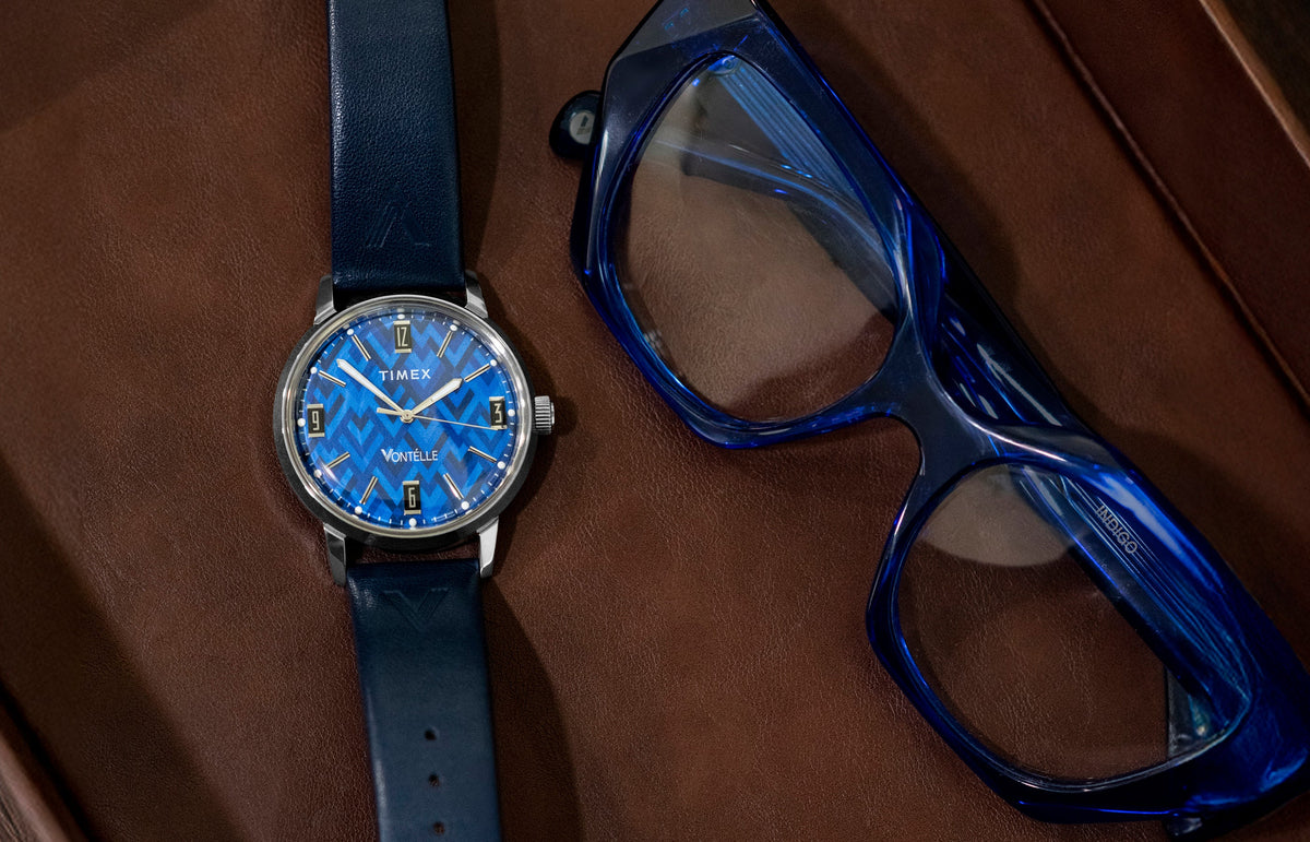 TW2Y73800 Blue patterned dial watch with a navy strap laid beside blue eyeglasses on a brown surface.