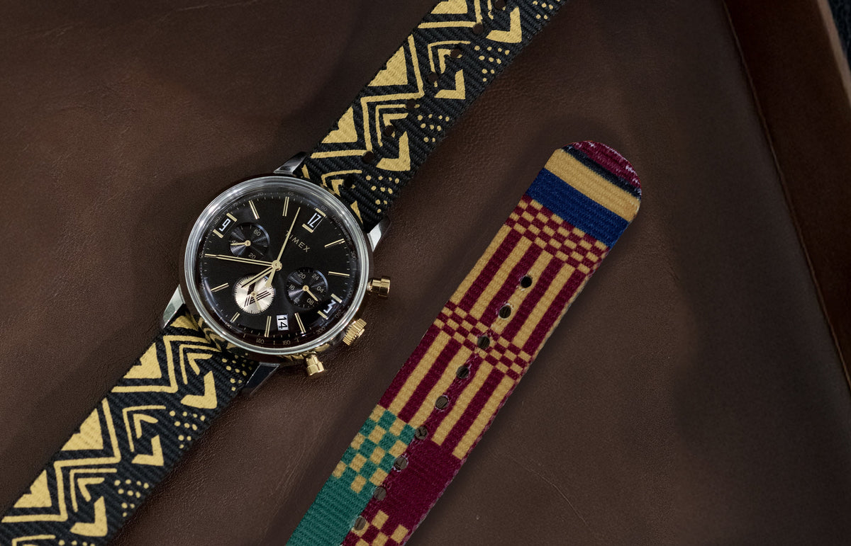 TWG069100 Vontélle x Timex Marlin Quartz Chronograph with a black dial and black-and-gold patterned strap laid beside a colorful striped strap on a brown surface.