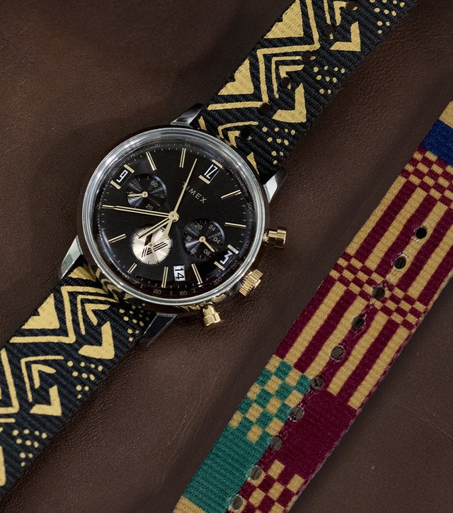 TWG069100 Vontélle x Timex Marlin Quartz Chronograph with a black dial and black-and-gold patterned strap laid beside a colorful striped strap on a brown surface.