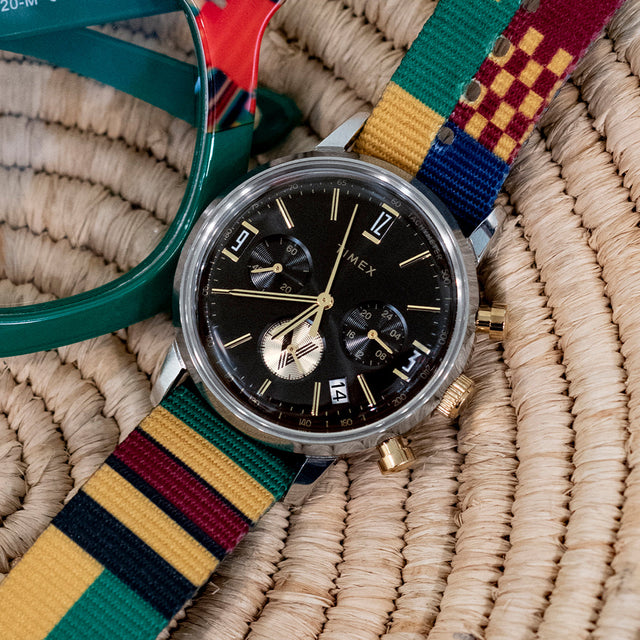 TWG069100 Watch with a black dial and colorful striped strap displayed on a woven surface beside green glasses.