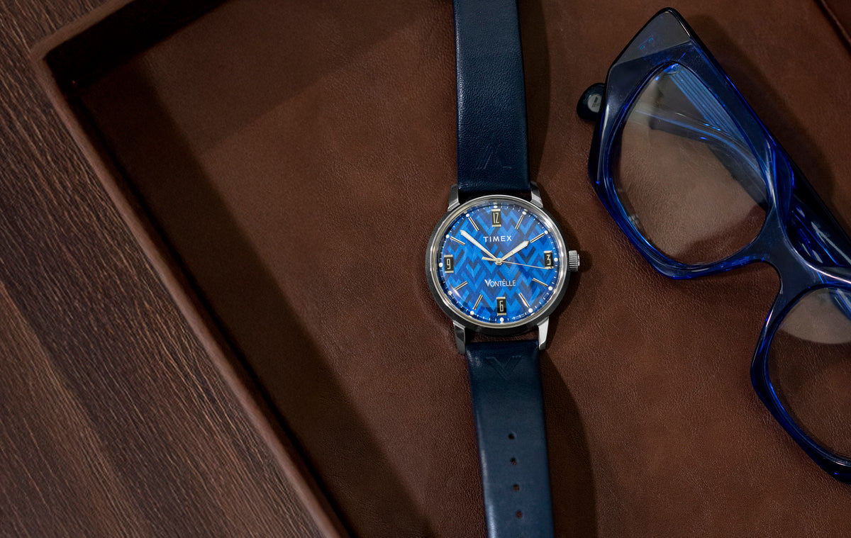 TW2Y73800 Vontélle x Timex Marlin watch with a blue geometric dial, silver-tone case and navy strap beside blue eyeglasses on a brown leather tray.