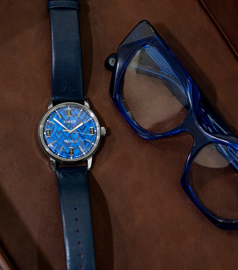 TW2Y73800 Vontélle x Timex Marlin watch with a blue geometric dial, silver-tone case and navy strap beside blue eyeglasses on a brown leather tray.