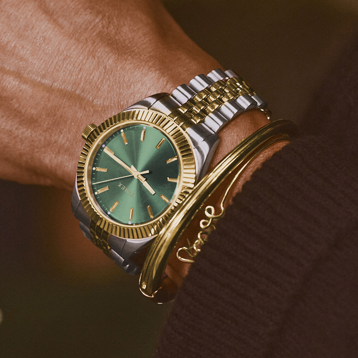 Two-tone Timex watch with a green dial worn alongside gold bracelets.