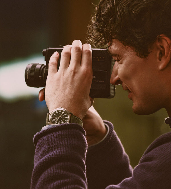 Gifts for Him - Man using a vintage movie camera while wearing a Timex watch with a black dial and green strap.