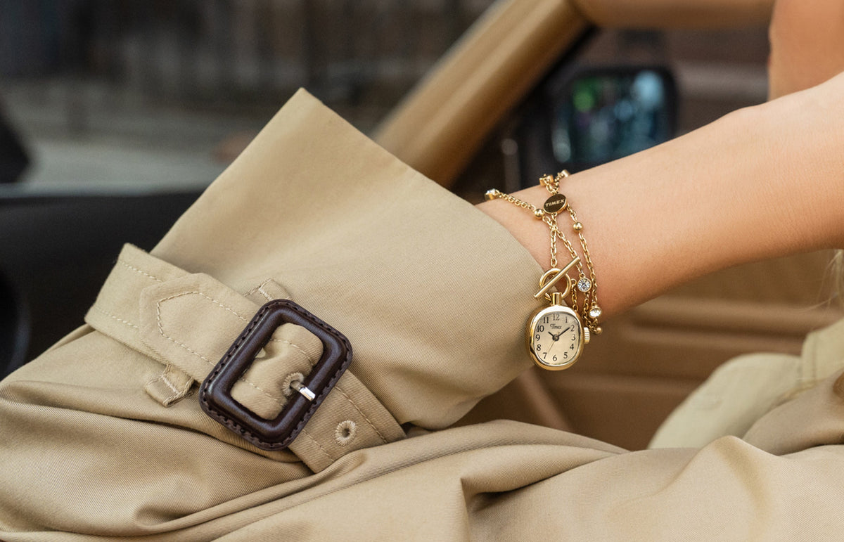 TW6A17700 Close-up of a gold-tone Cavatina® Mini watch necklace wrapped around a wrist beside a beige trench coat cuff.