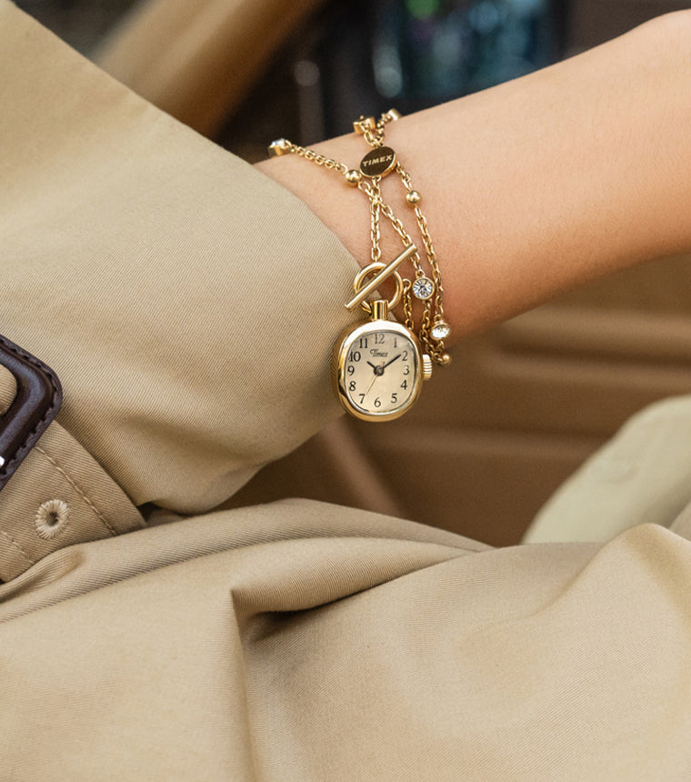 TW6A17700 Close-up of a gold-tone Cavatina® Mini watch necklace wrapped around a wrist beside a beige trench coat cuff.