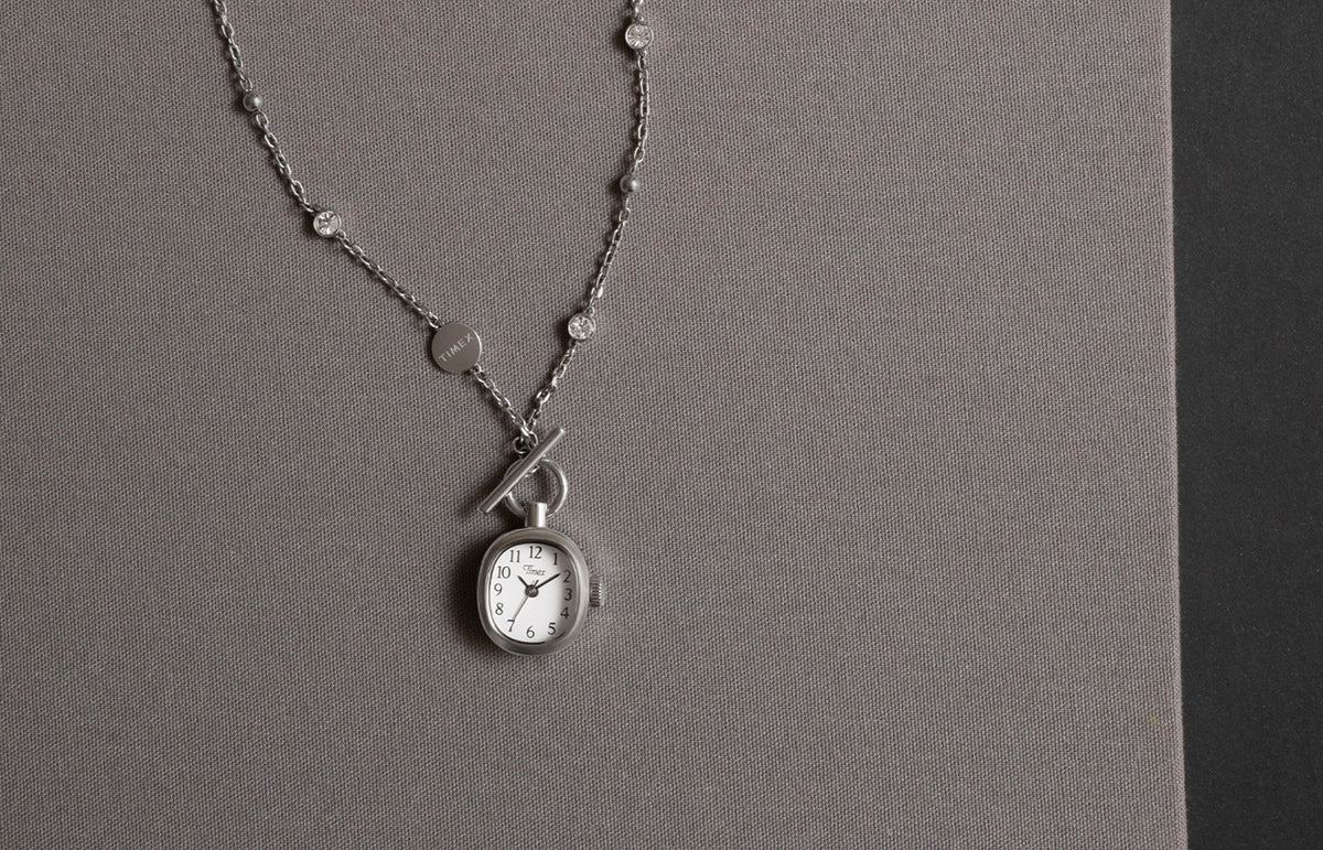 TW6A17600 Cavatina® Mini necklace with an oval white-dial watch pendant and silver-tone chain with clear stones, displayed on gray fabric.
