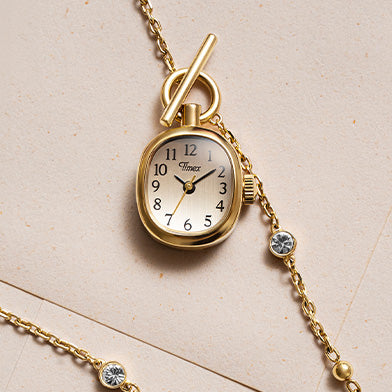 TW6A17700 Cavatina® Mini gold-tone watch pendant with black Arabic numerals, beside a chain with clear stones on cream paper.