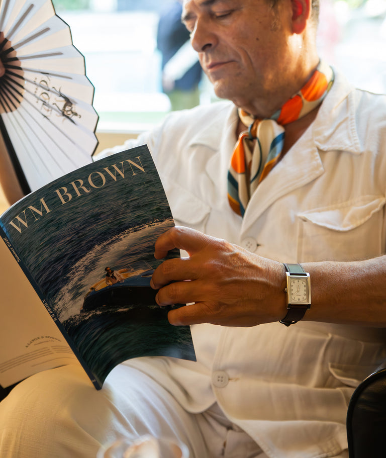 TWG069000 Man reading WM Brown magazine and holding a folding fan while wearing a rectangular WM Brown x Timex watch with a cream dial and dark leather strap.