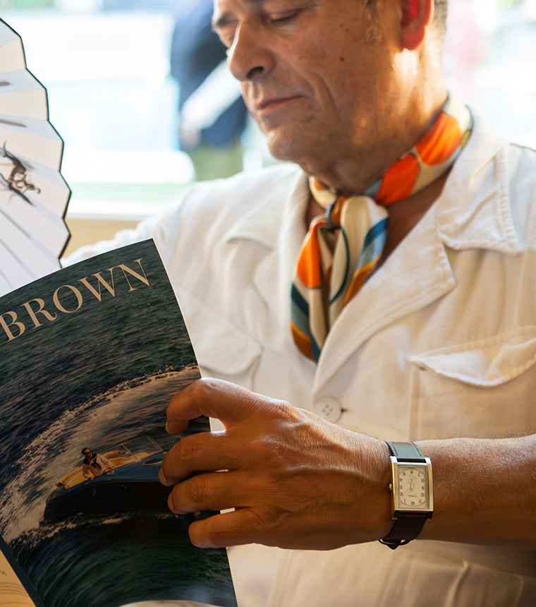TWG069000 Man reading WM Brown magazine and holding a folding fan while wearing a rectangular WM Brown x Timex watch with a cream dial and dark leather strap.