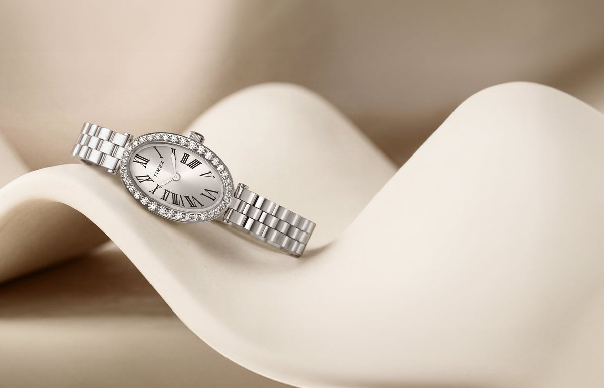 TW2Y90000 Timex Cavatina® Crystals watch with an oval Roman numeral dial, crystal-lined bezel, and silver-tone bracelet, resting on a curved ivory display.