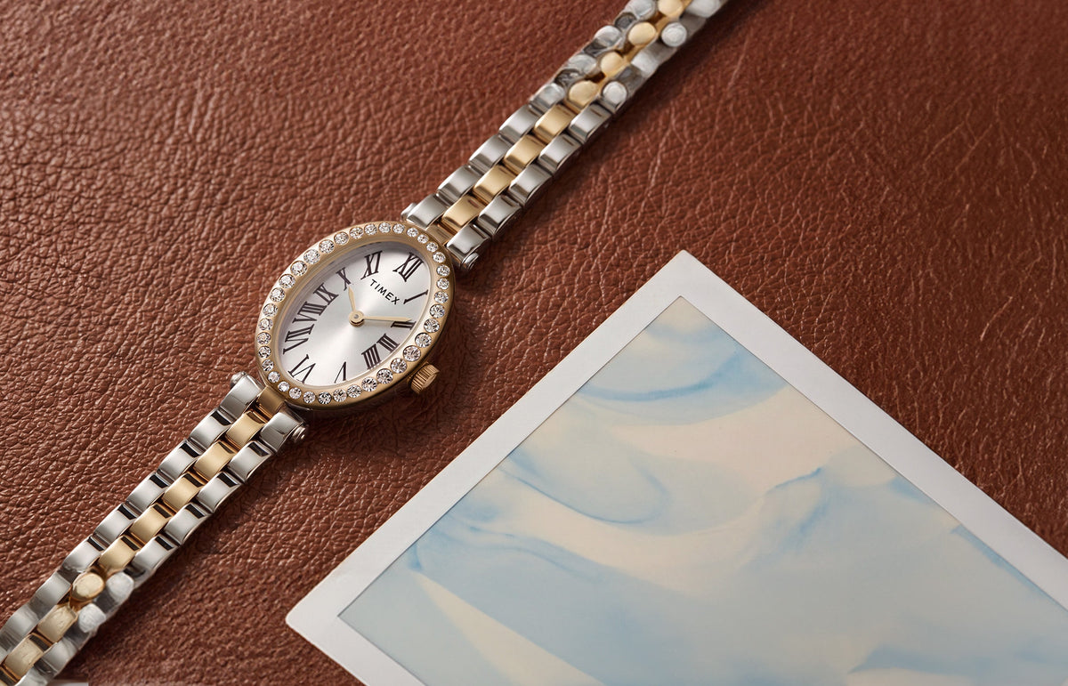 TW2Y90200 Timex Cavatina® Crystals watch with a two-tone bracelet, silver-tone oval dial, and crystal-lined gold-tone bezel on brown leather beside a photograph.