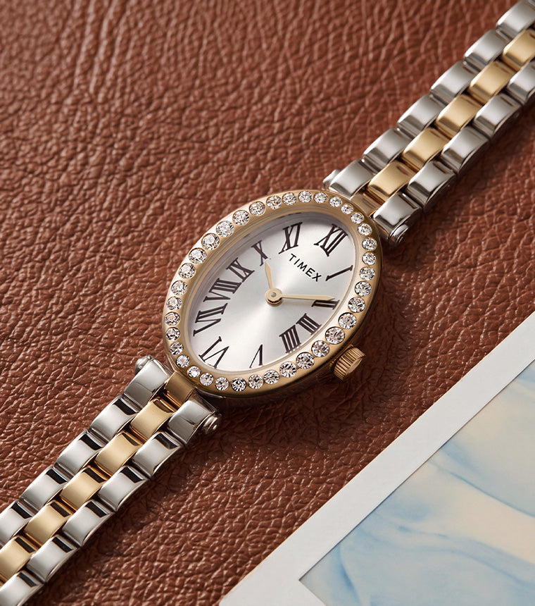 TW2Y90200 Timex Cavatina® Crystals watch with a two-tone bracelet, silver-tone oval dial, and crystal-lined gold-tone bezel on brown leather beside a photograph.