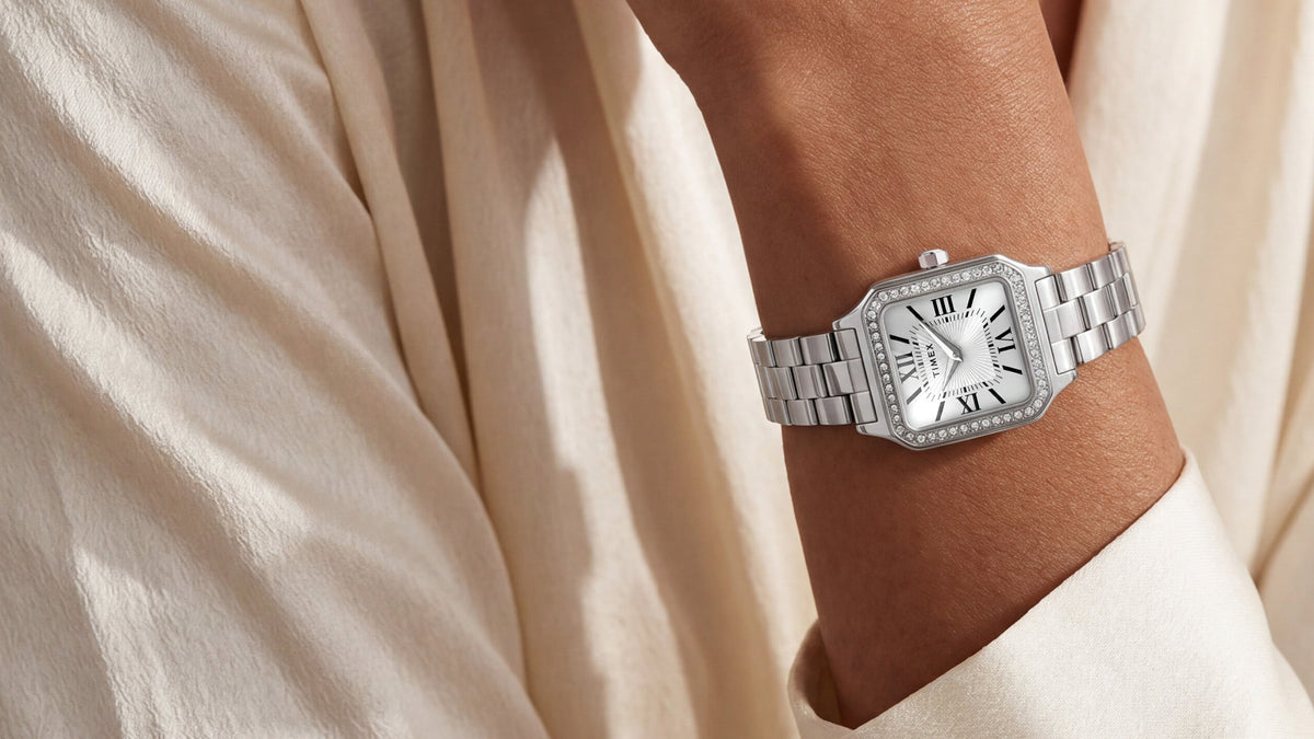 TW2Y90300 Silver-tone Timex Addison watch with a white Roman numeral dial and crystal-lined bezel, worn on a wrist against an ivory blouse.