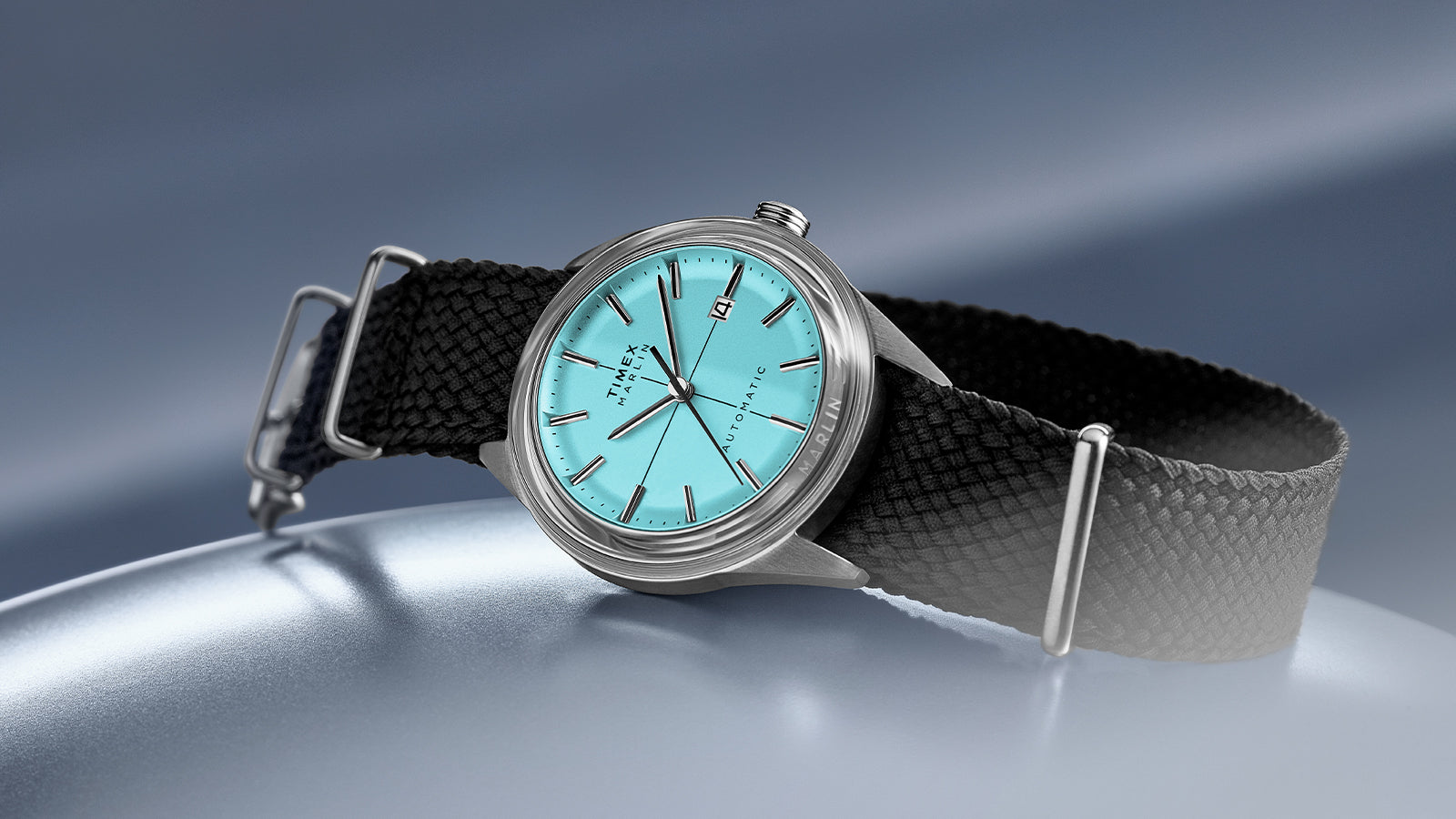 Timex Marlin Jet Automatic with aqua blue dial, stainless steel case, and black woven strap displayed on a metallic blue surface.
