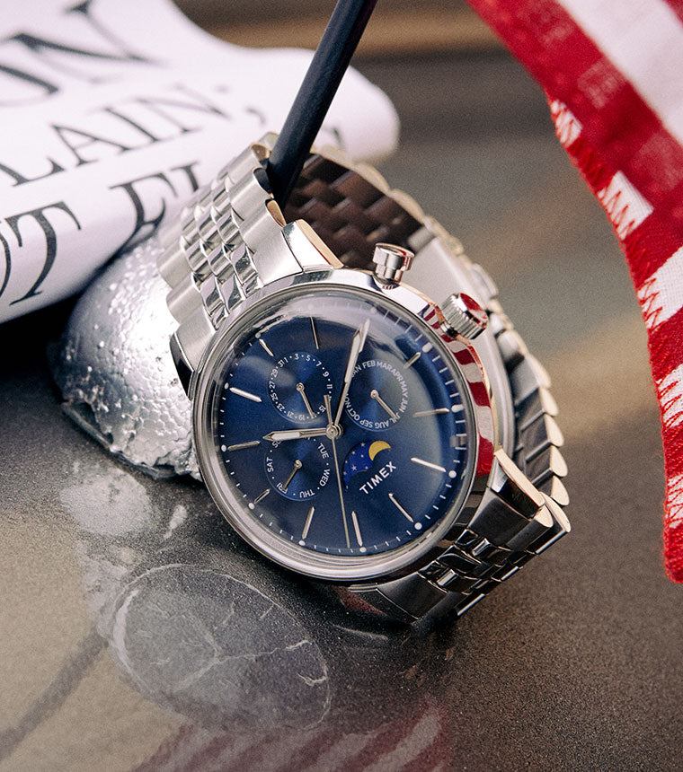 Image features our marlin moon phase watch which has a blue dial and a silver tone bracelet.  The watch is leaning on a half moon which is holding the American Flag