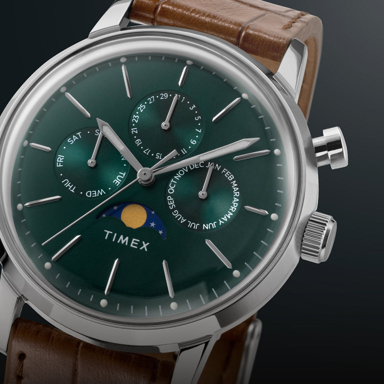 Close-up of the Timex Marlin® Moon Phase watch featuring a deep green dial with silver-tone baton hour markers and hands, three subdials displaying the day, date, and month, a moon phase display at 6 o'clock, a polished stainless steel case, and a brown leather strap.