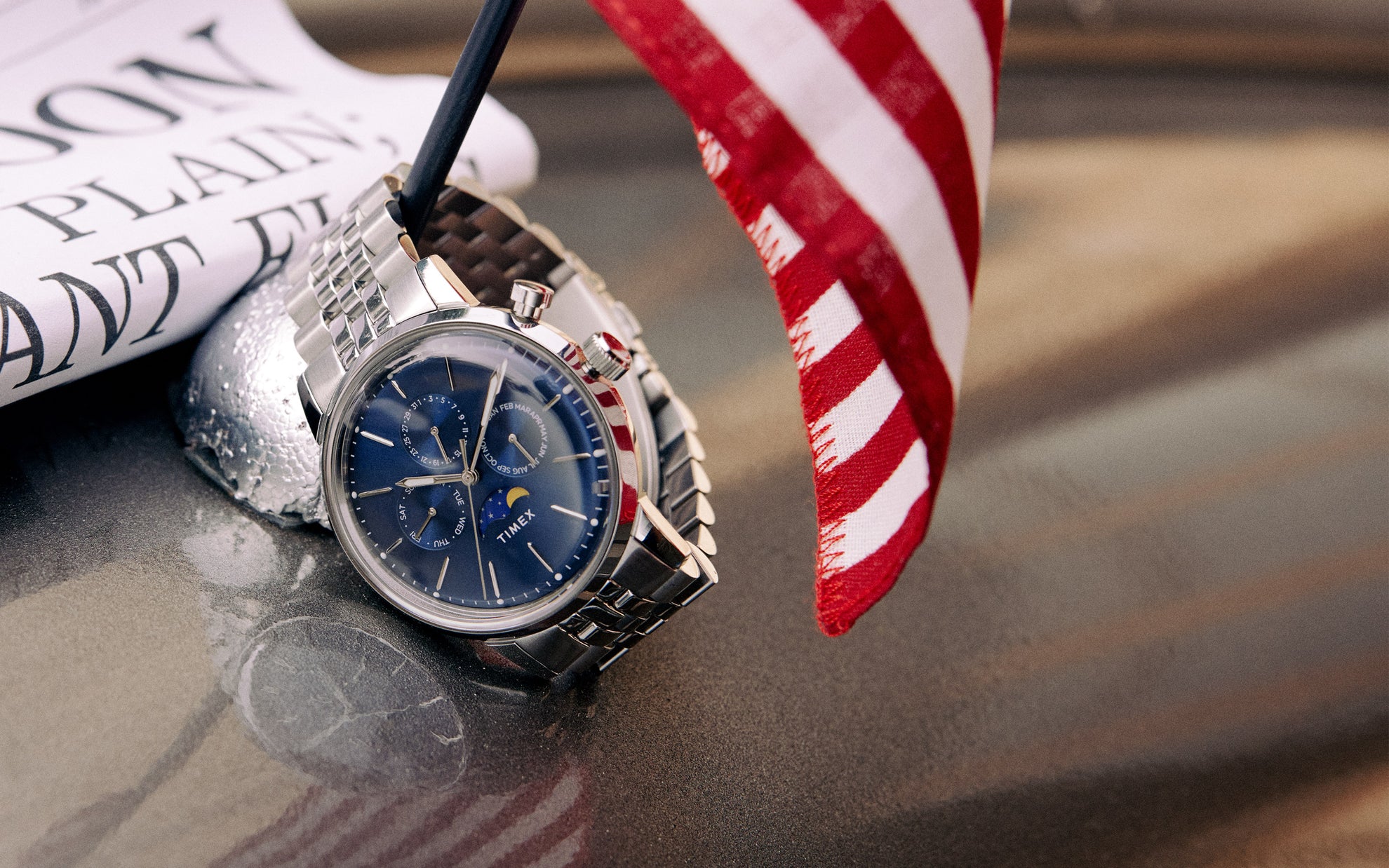 Image features our marlin moon phase watch which has a blue dial and a silver tone bracelet.  The watch is leaning on a half moon which is holding the American Flag
