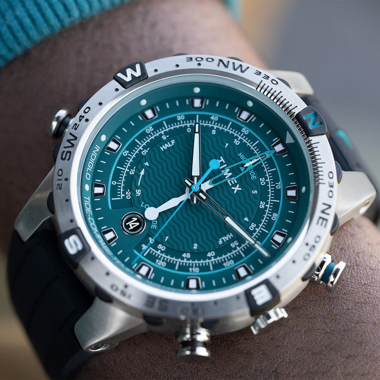A Timex wristwatch with a teal dial featuring wave-pattern detailing, luminous hands, and a date window at 6 o’clock. The stainless steel case includes a compass bezel marked with cardinal directions, paired with a black textured strap. The watch is worn on the wrist, highlighting its sporty and functional design.