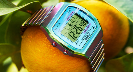Digital watch with expandable strap in shades of purple and blue. Watch is lying across oranges. 