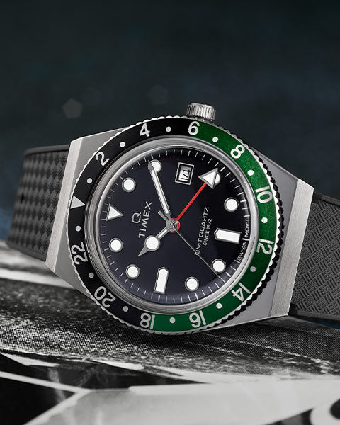 A Q Timex wristwatch with a black dial, luminous hour markers, and a rotating two-tone bezel in black and green. The watch features a date window at the 3 o’clock position, a stainless steel case, and a textured black rubber strap. The background is dark and blurred, emphasizing the watch's sleek and sporty design.