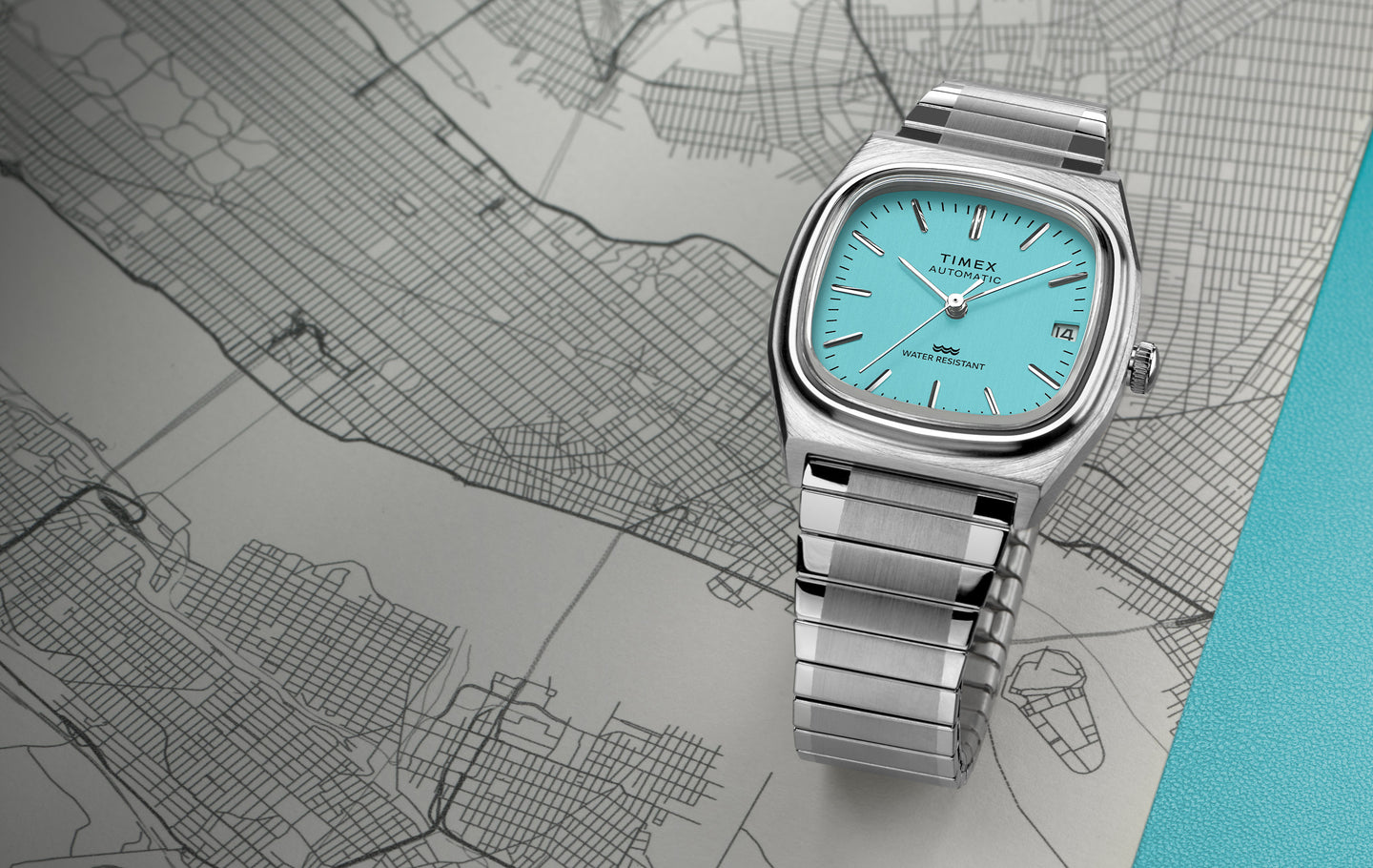 Timex® Automatic 1983 E Line watch with a turquoise dial and stainless-steel bracelet resting on a city map.