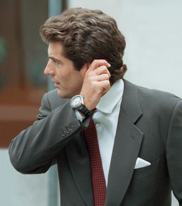 A man in a business suit adjusting his hair with his right hand, wearing a Intrepid Reissue, JFK watch on his left wrist.