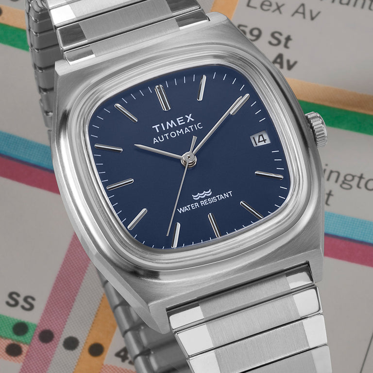 A close-up of a Timex E-Line Automatic wristwatch with a deep blue dial, minimalist silver hour markers, and a date display. The watch features a sleek, brushed stainless steel case and bracelet, evoking a refined and modern aesthetic. It is set against a background of a New York City subway map, symbolizing urban style and functionality.