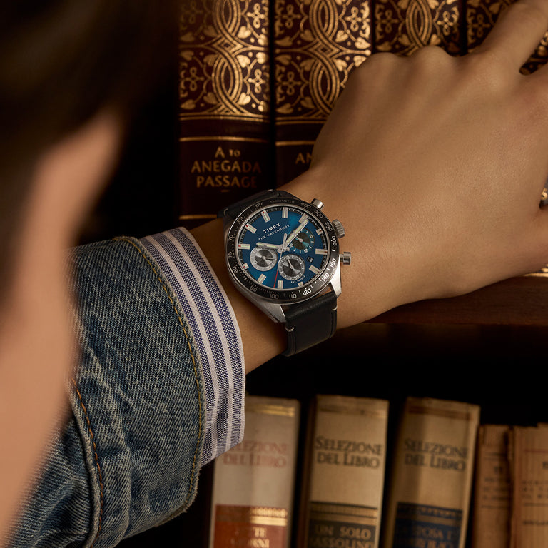 Timex Waterbury chronograph watch with a blue dial, subdials, tachymeter bezel, and black leather strap, worn on a wrist in front of a bookshelf of vintage books.