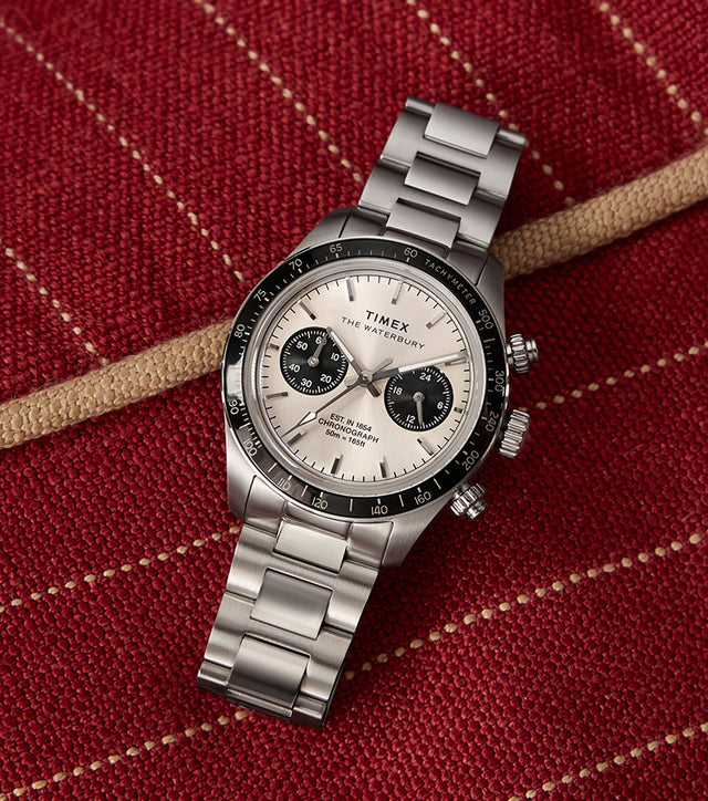 A detailed shot of the Timex Waterbury Heritage Chronograph watch resting on a rich, textured red fabric with beige stitching. The stainless steel bracelet, black tachymeter bezel, and silver dial with contrasting black subdials are prominently displayed, highlighting the watch’s refined yet sporty design.