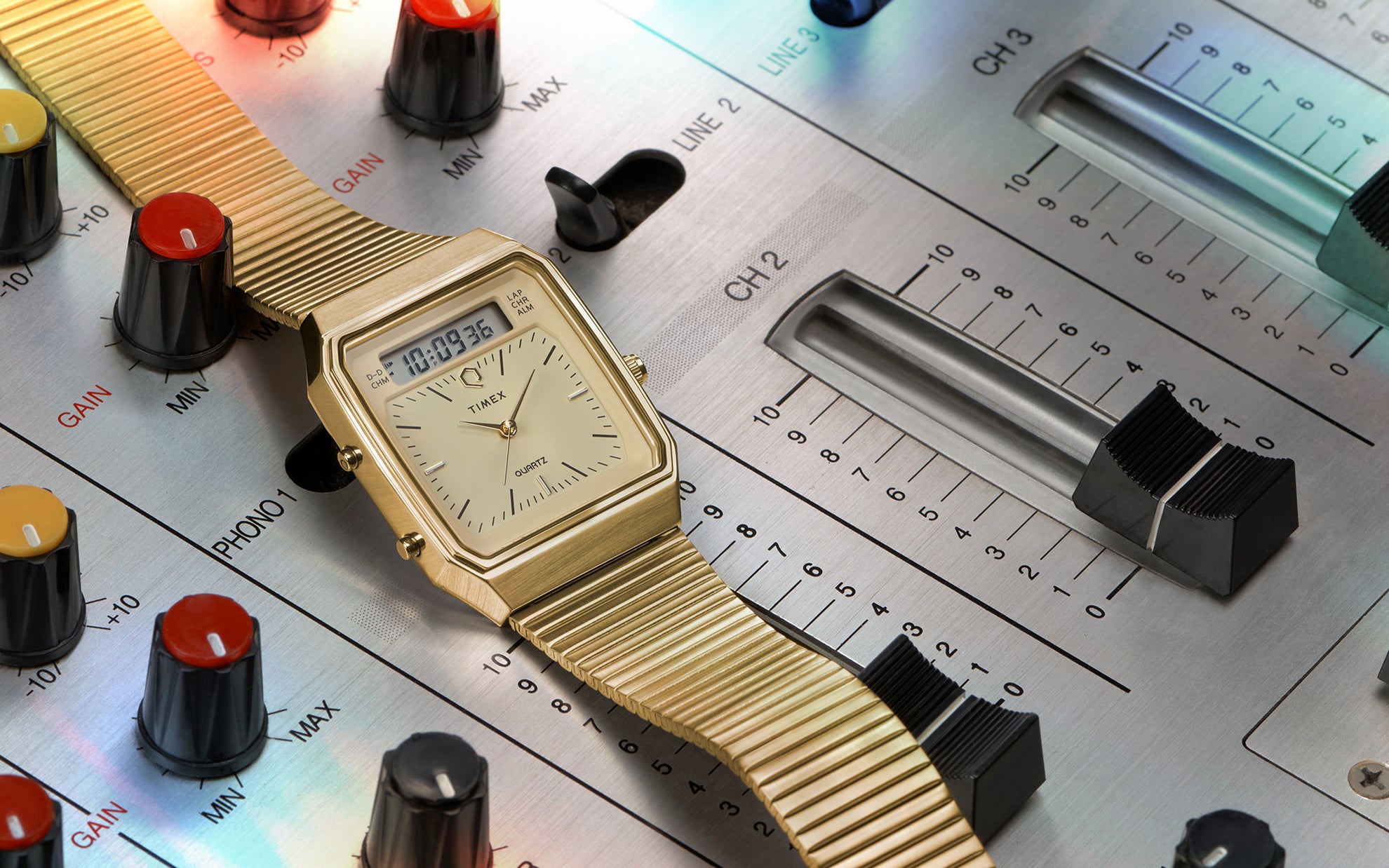 A gold-tone Q Timex® 1982 Ana-Digi Reissue watch placed on a vintage audio mixing console. The watch features a champagne-colored dial with an analog and digital hybrid display, a rectangular case, and a flexible stainless steel bracelet. The background showcases a professional DJ mixer with colorful knobs and sliders, enhancing the retro aesthetic of the timepiece.