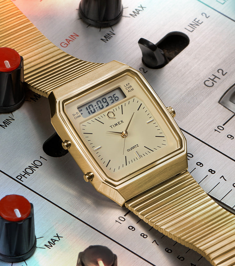 A gold-tone Q Timex® 1982 Ana-Digi Reissue watch placed on a vintage audio mixing console. The watch features a champagne-colored dial with an analog and digital hybrid display, a rectangular case, and a flexible stainless steel bracelet. The background showcases a professional DJ mixer with colorful knobs and sliders, enhancing the retro aesthetic of the timepiece.