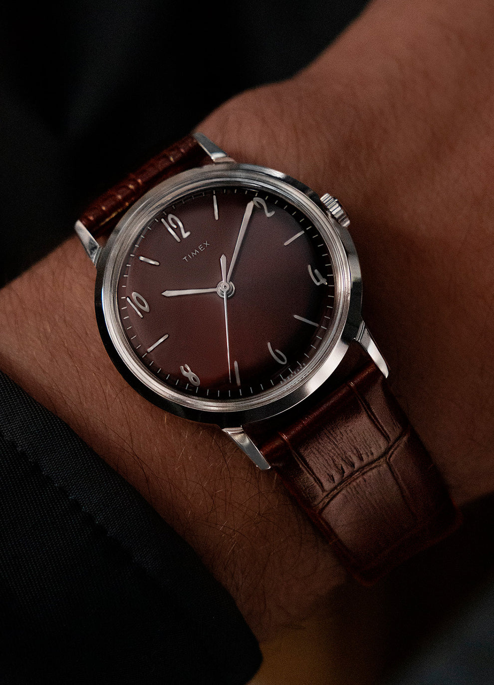 A Timex wristwatch with a brown leather strap and a brown dial, worn on a person's wrist, dressed in a dark suit.