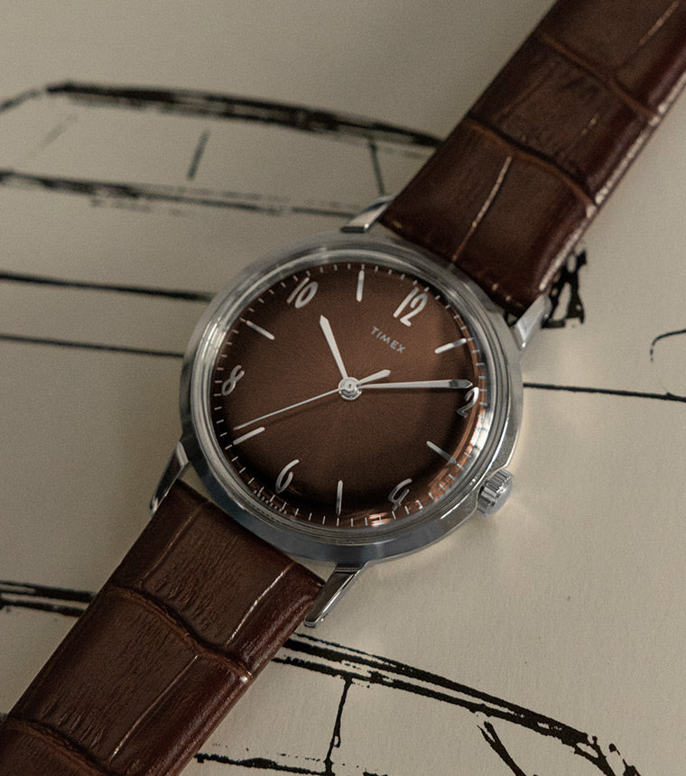 A Timex wristwatch with a brown leather strap and a deep brown dial, placed on a vintage-style sketch of a car with a handwritten signature.